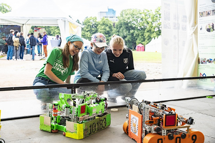 Drei Kinder beobachten gespannt zwei kleine Roboter in einer Arena und verfolgen deren Bewegungen. Die Szene zeigt ein Robotik-Angebot bei Explore Science im Herzogenriedpark Mannheim und vermittelt die Begeisterung junger Besucherinnen und Besucher für Technik und Wissenschaft.